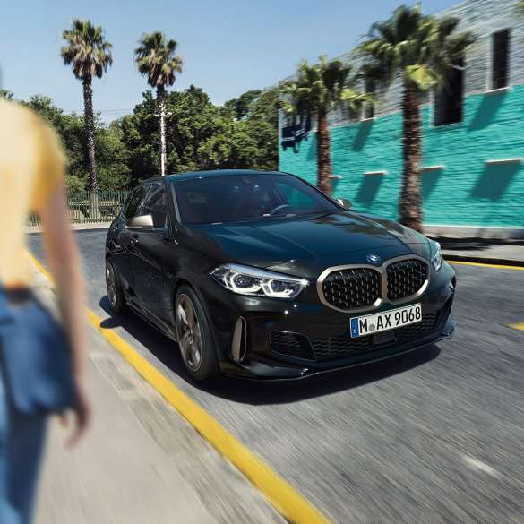 BMW M135i xDrive, вид спереду з поворотом в три чверті. BMW M135i xDrive в Black Sapphire металік, вид спереду за кермом з поворотом в три чверті з жінкою на тлі.