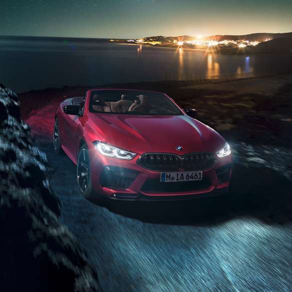 BMW M8 Competition Кабріолет, вид спереду з поворотом в три чверті. BMW M8 Competition Кабріолет в Motegi Red металік з відкритим м'яким верхом, вид спереду з поворотом в три чверті, їзда вночі по прибережній дорозі.