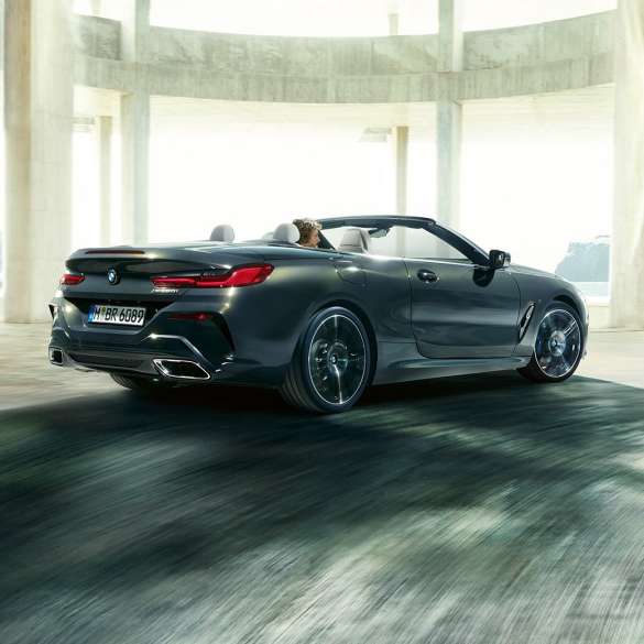 BMW M850i xDrive Кабріолет, вид ззаду з поворотом в три чверті. BMW M850i xDrive Кабріолет в Dravit Grey металік з відкритим м'яким верхом, вид ззаду з поворотом в три чверті, перед бетонним будівлею.