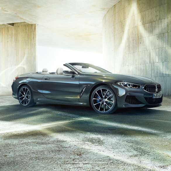 BMW M850i xDrive Кабріолет, вид збоку з поворотом в три чверті. BMW M850i xDrive Кабріолет в Dravit Grey металік з відкритим м'яким верхом, вид збоку з поворотом в три чверті, перед бетонним будівлею.