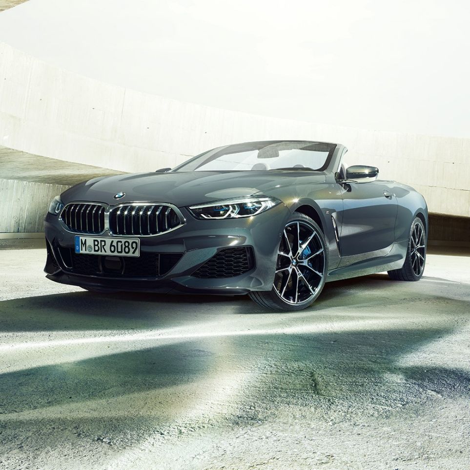 BMW M850i xDrive Кабріолет, вид спереду з поворотом в три чверті. BMW M850i xDrive Кабріолет в Dravit Grey металік з відкритим м'яким верхом, вид спереду з поворотом в три чверті, перед бетонним будівлею.
