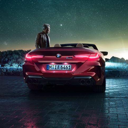 BMW M8 Competition Кабріолет, вид ззаду. BMW M8 Competition Кабріолет в Motegi Red металік з відкритим м'яким верхом, вид ззаду, з чоловіком під зоряним небом.
