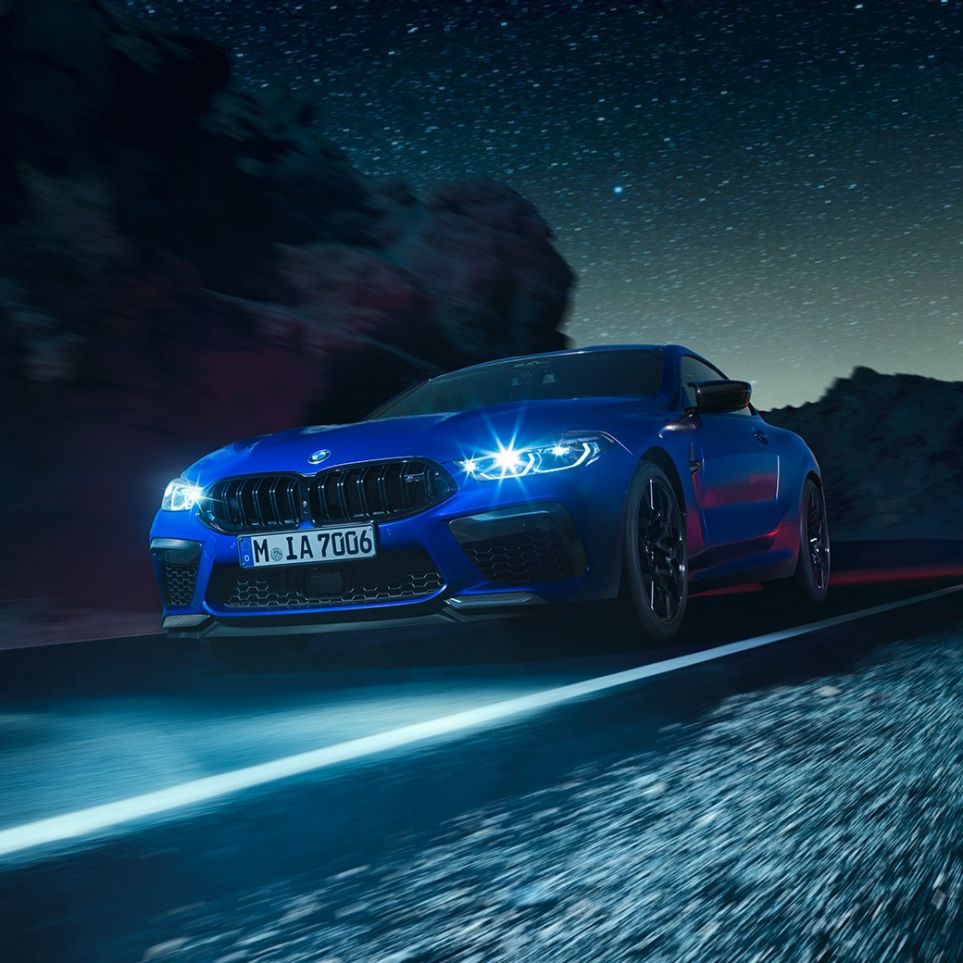 BMW M8 Competition Coupé, вид спереду з поворотом в три чверті. BMW M8 Competition Coupé в Marina Bay Blue металік з включеними світлодіодними фарами, вид спереду з поворотом в три чверті, водіння вночі.