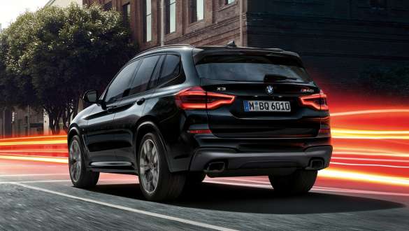 BMW X3 M40i, вид ззаду з поворотом в три чверті. BMW X3 M40i в Black Sapphire металік, за кермом, екстер'єр, вид ззаду з поворотом в три чверті на міському тлі зі світловими смужками.
