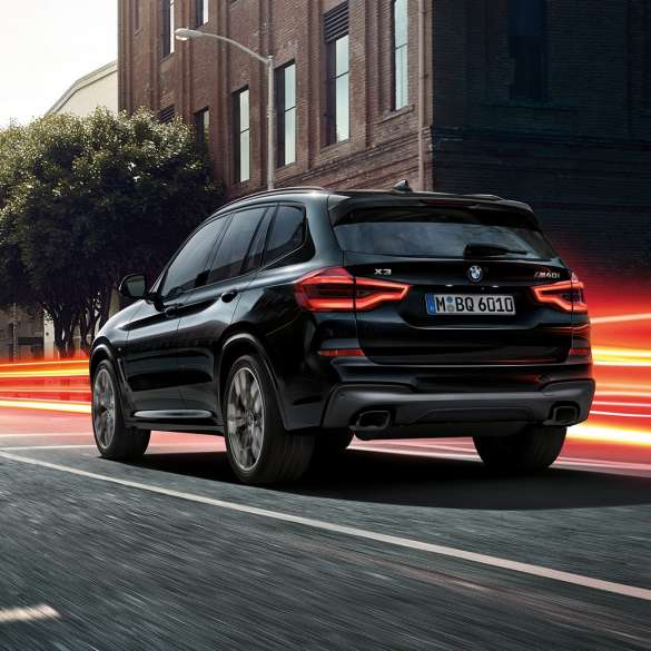 BMW X3 M40i в Black Sapphire, вид ззаду з поворотом в три чверті. BMW X3 M40i в Black Sapphire металік, за кермом, екстер'єр, вид ззаду з поворотом в три чверті на міському тлі зі світловими смужками.