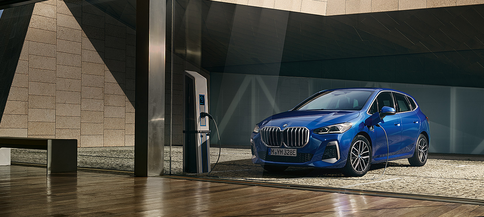 BMW 2 серії Active Tourer 230e плагін-гібрид U06 2021 M у кольорі Portimao Blue металік у три чверті на зарядній станції