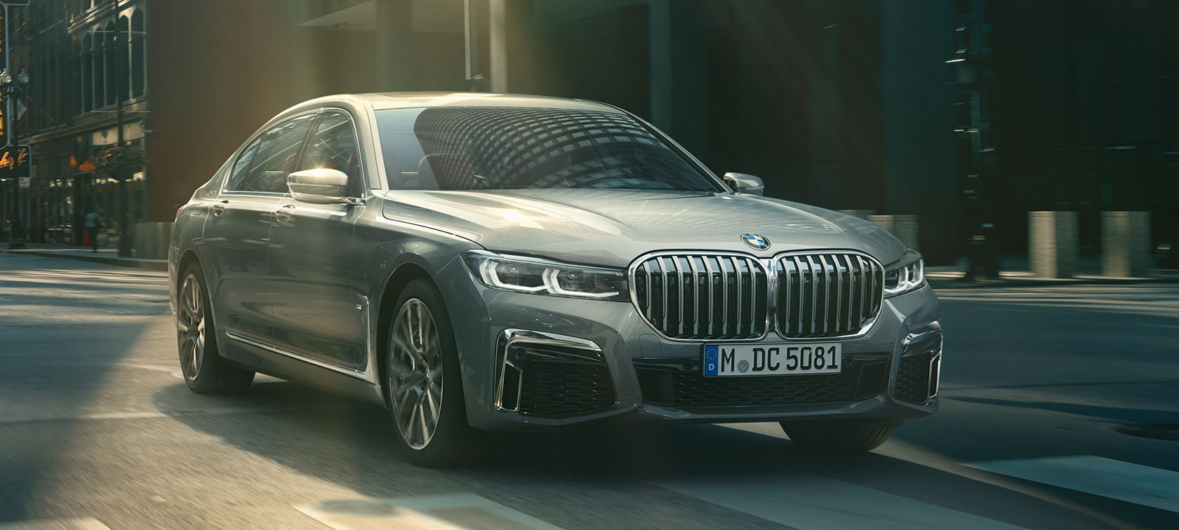 BMW 7 серії Седан сірого кольору в русі по міських вулицях: вид спереду з поворотом в три чверті BMW 7 серії Седан сірого кольору в русі: вид спереду з поворотом в три чверті