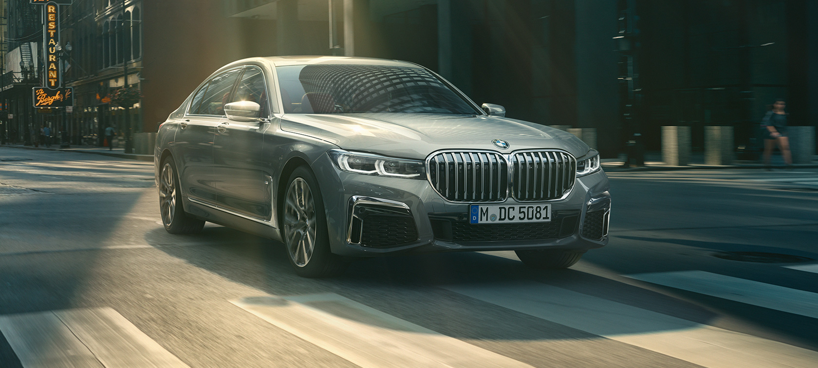 BMW 7 серии Седан G11 G12 2019 в цвете Bernina Grey Amber Effect, вид спереди на три четверти в движении BMW 7 серии Седан G11 G12 2019 в цвете Bernina Grey Amber Effect, вид спереди на три четверти при езде в городе