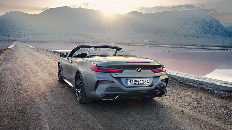 BMW 8 серії Cabrio G14 LCI Facelift 2022 у кольорі Individual Frozen Pure Grey metallic, вид ззаду з відкритим дахом на пісчаній дорозі з пагорбами на фоні