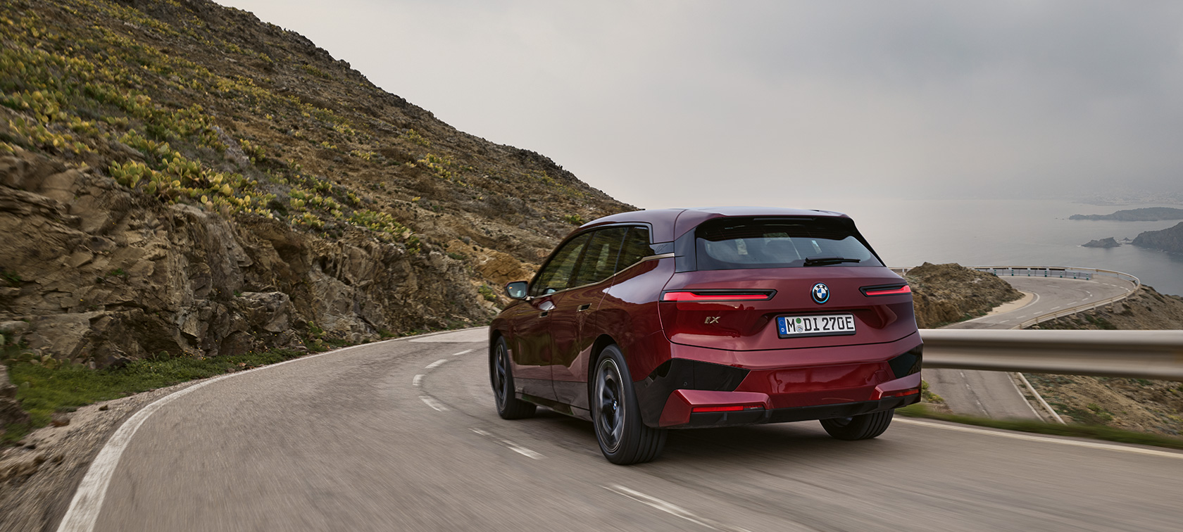 BMW iX i20 2021 електричний кросовер BMW iX xDrive50 під час руху узбережжям BMW iX i20 2021 електричний кросовер BMW iX xDrive50 кольору Aventurin Red вид ззаду під час руху узбережжям