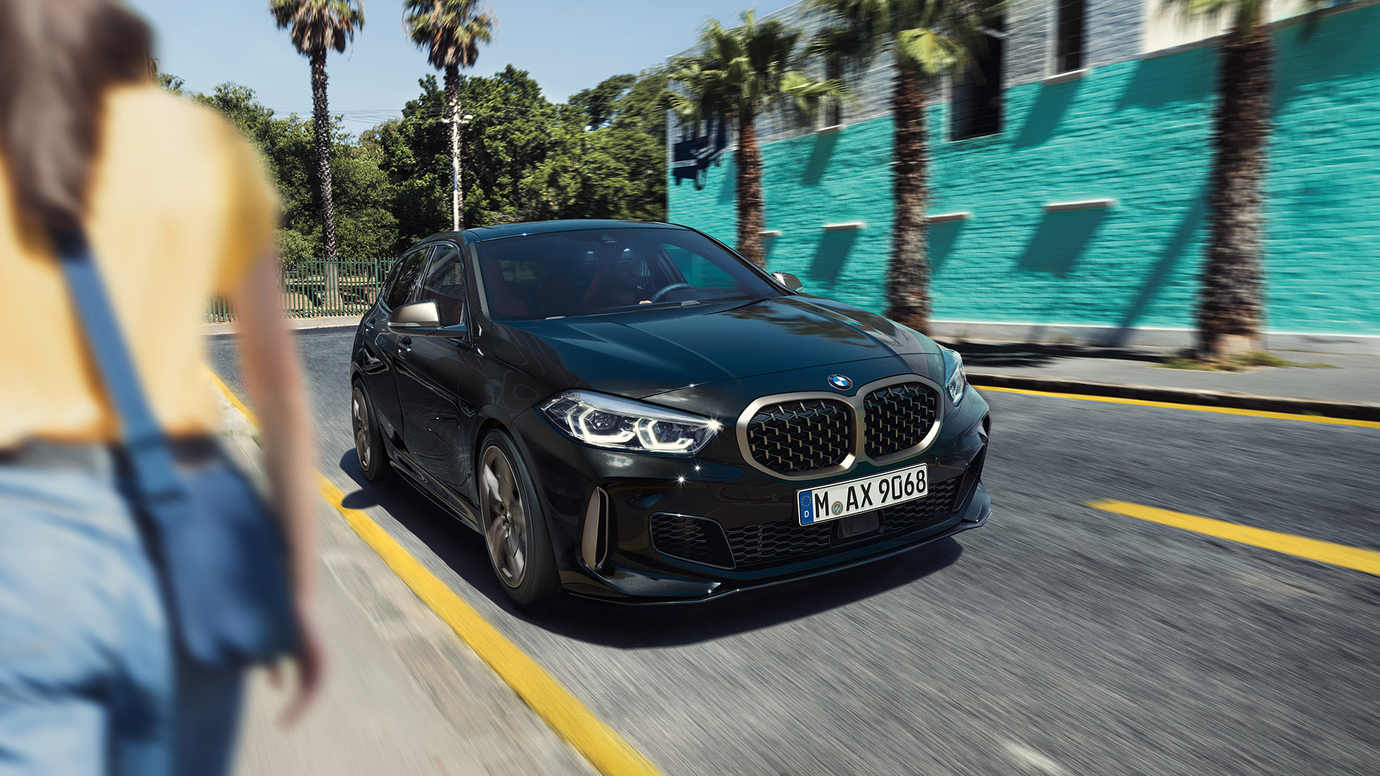 BMW M135i xDrive, вид спереду з поворотом в три чверті. BMW M135i xDrive в Black Sapphire металік, вид спереду за кермом з поворотом в три чверті з жінкою на тлі.
