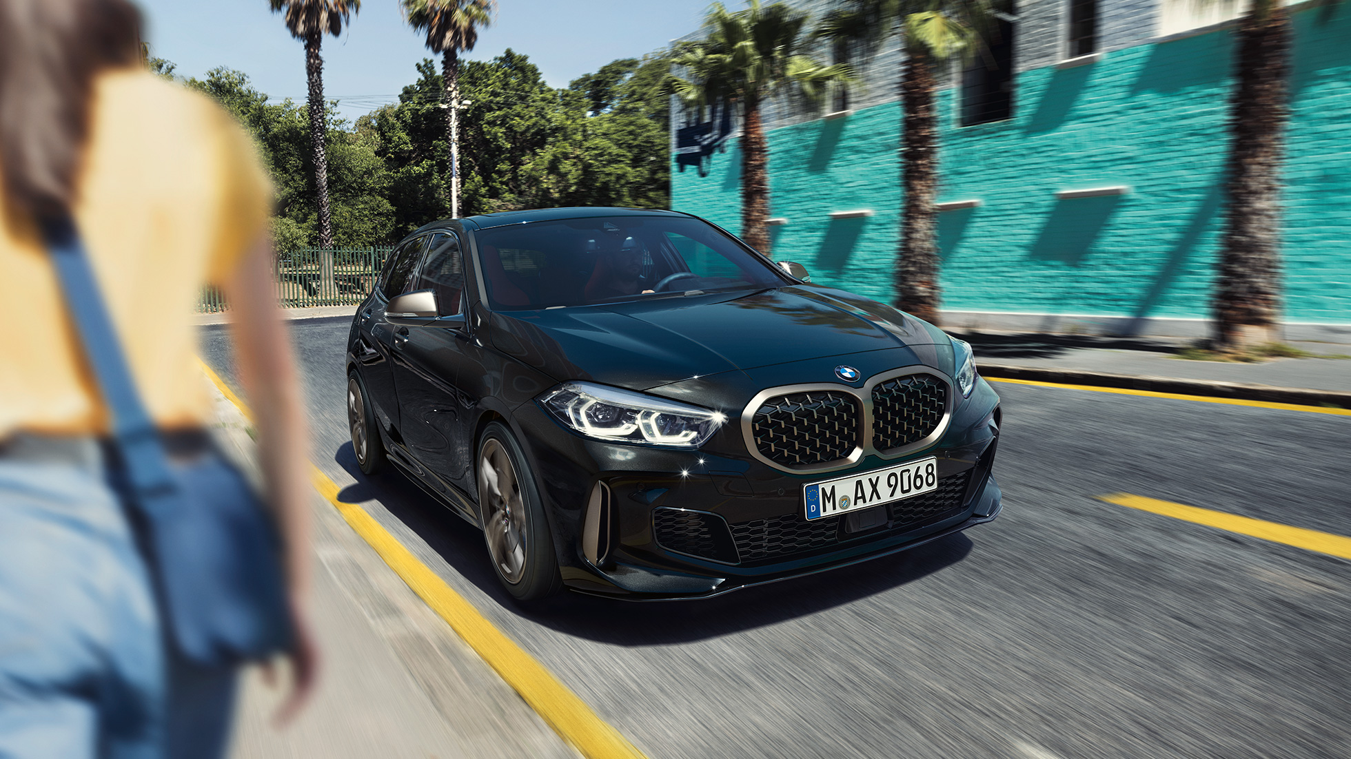 BMW M135i xDrive, вид спереду з поворотом в три чверті. BMW M135i xDrive в Black Sapphire металік, вид спереду за кермом з поворотом в три чверті з жінкою на тлі.