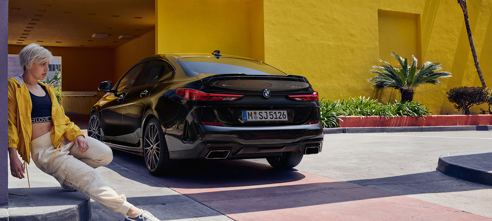 BMW M235i xDrive Gran Coupé вид ззаду з поворотом в три чверті BMW M235i xDrive Gran Coupé Black Sapphire металік F44 вид ззаду з поворотом в три чверті з жінкою попереду