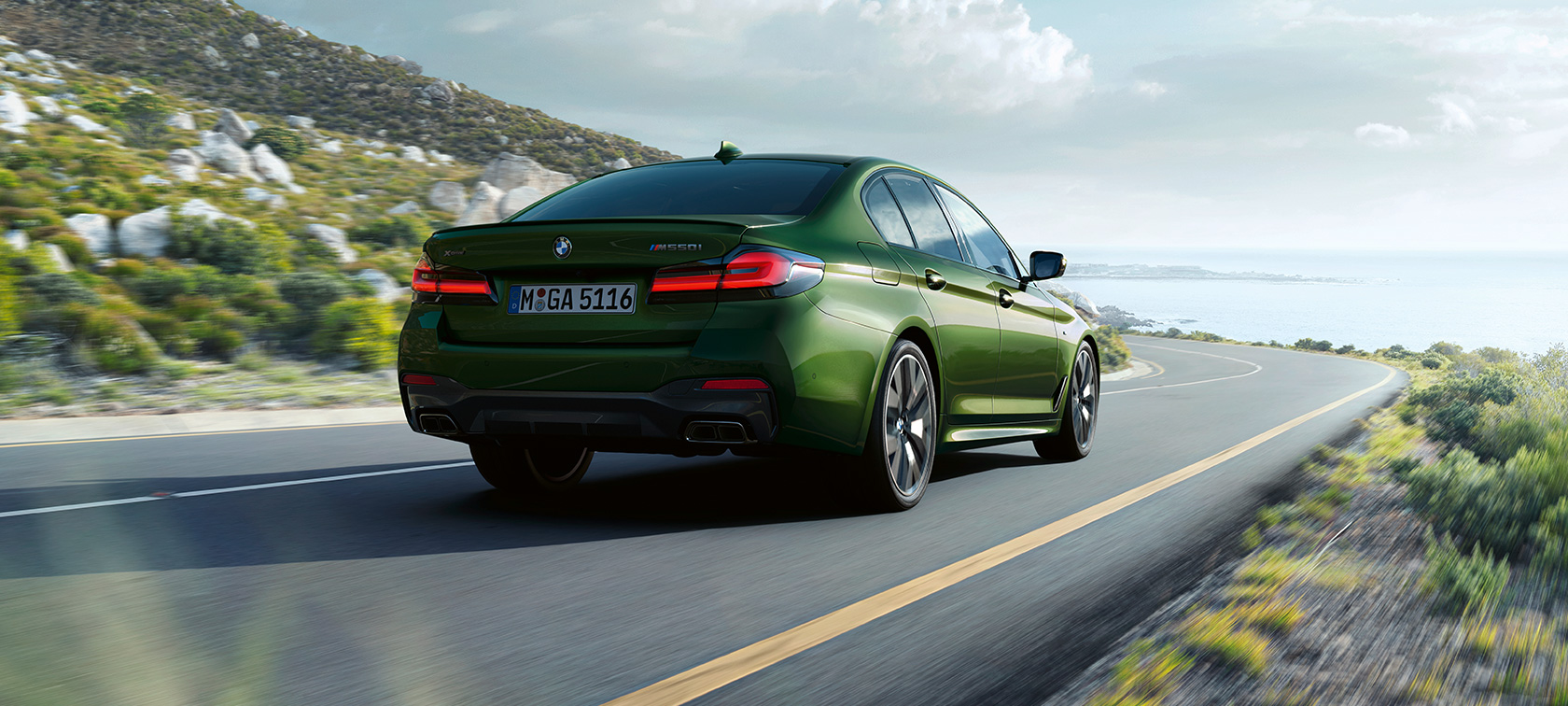 BMW M550i xDrive Седан G30 LCI 2020 вид на три чверті ззаду під час руху BMW M550i xDrive Седан G30 LCI 2020 кольору BMW Individual Verde Ermes Metallic вид на три чверті ззаду під час руху центром міста