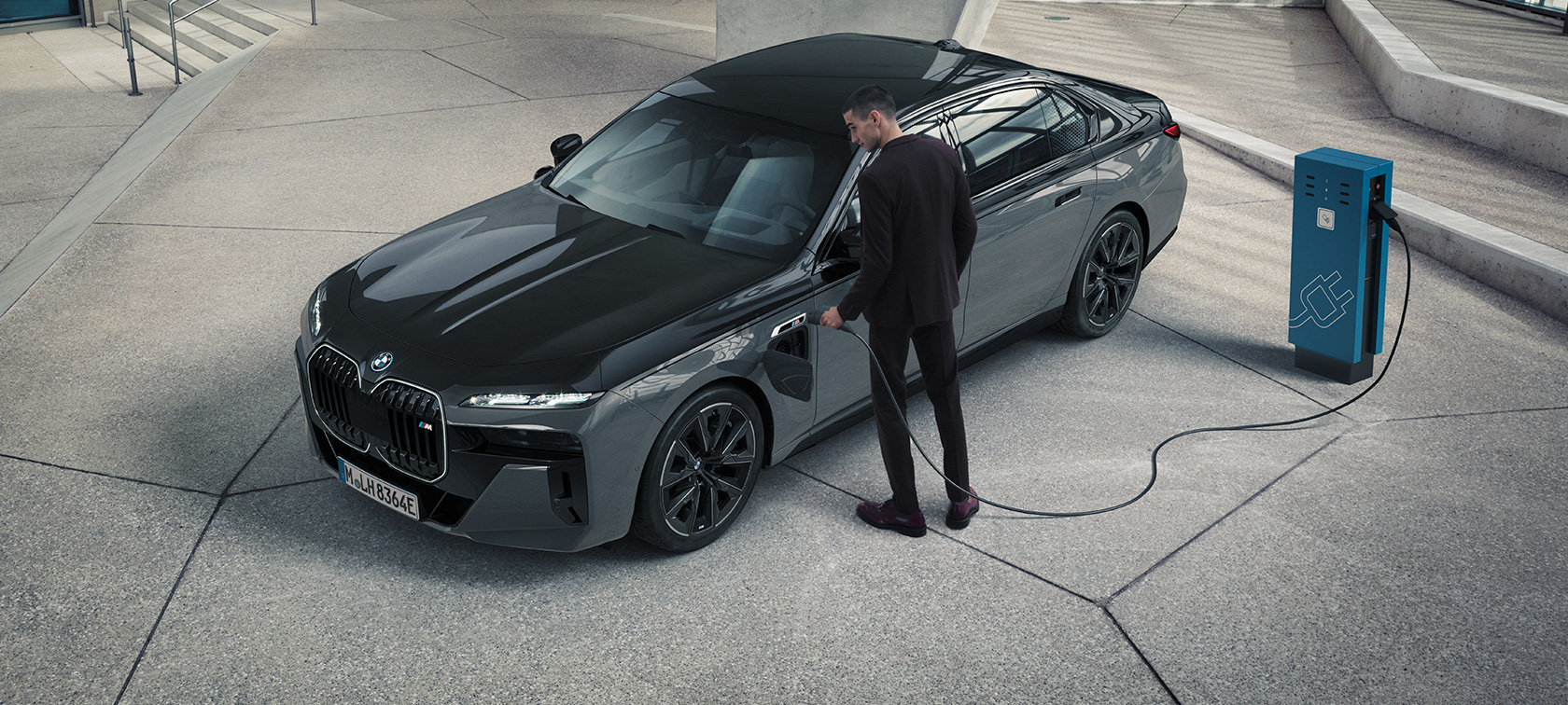 BMW M760e xDrive Седан G70 Пакет M Performance BMW Individual двокольорове забарвлення Black Sapphire металік BMW Individual Dravite Grey металік вид спереду в три чверті чоловік із зарядним кабелем поруч із зарядною станцією