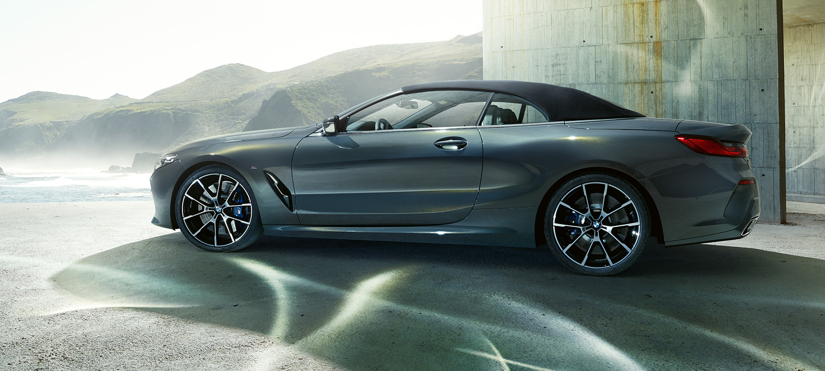 BMW M850i xDrive Кабріолет, вид збоку. BMW M850i xDrive Кабріолет в кольорі Dravit Grey металік з закритим м'яким верхом, вид збоку перед пагорбами.