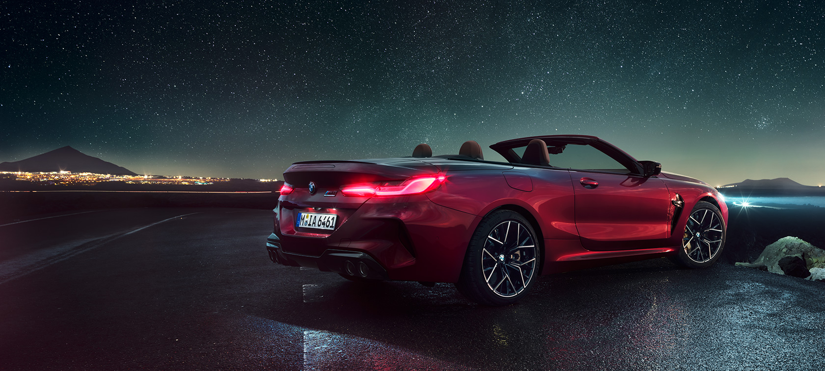 BMW M8 Competition Кабріолет, вид ззаду з поворотом в три чверті. BMW M8 Competition Кабріолет в Motegi Red металік з відкритим м'яким верхом, вид ззаду з поворотом в три чверті, під зоряним небом.