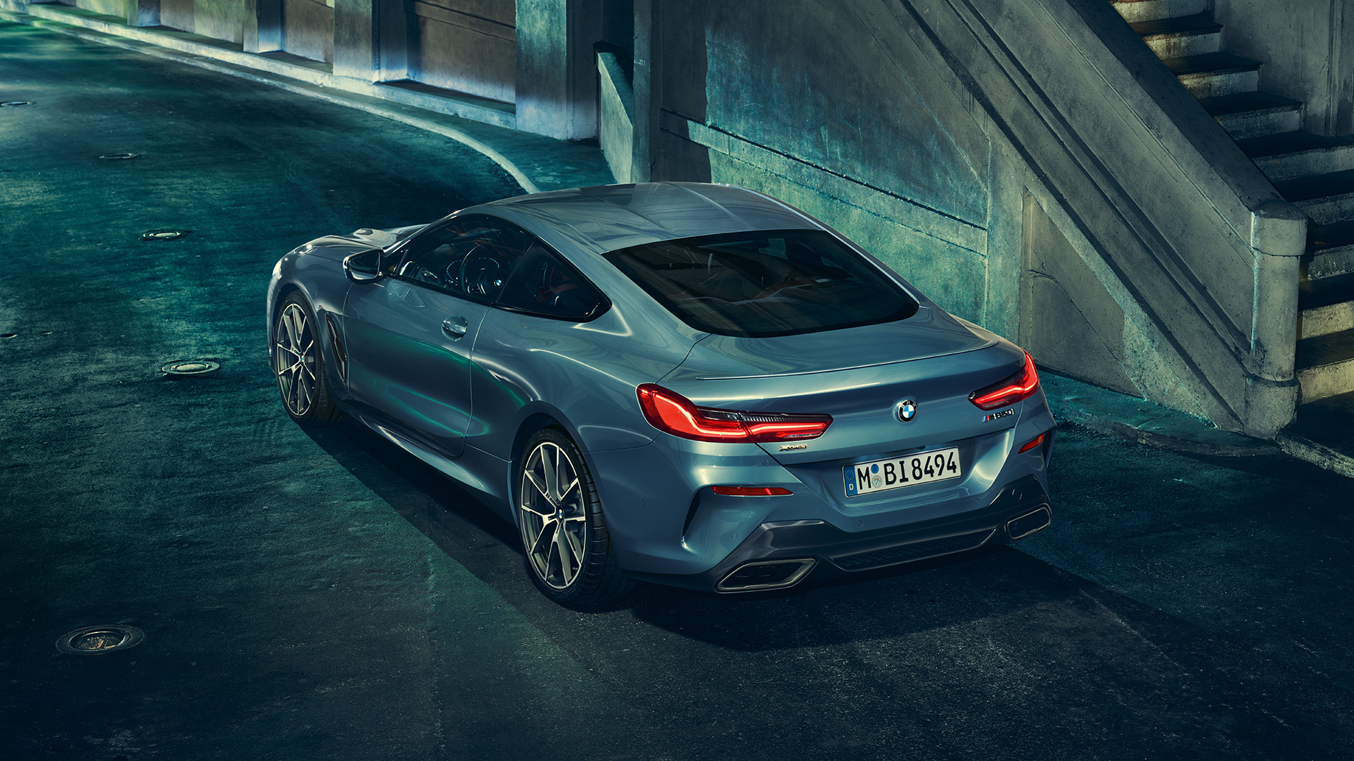 BMW M850i xDrive Coupé, вид ззаду з поворотом в три чверті зліва. BMW M850i xDrive Coupé в Barcelona Blue металік, вид ззаду з поворотом в три чверті ліворуч, стоїть вночі.