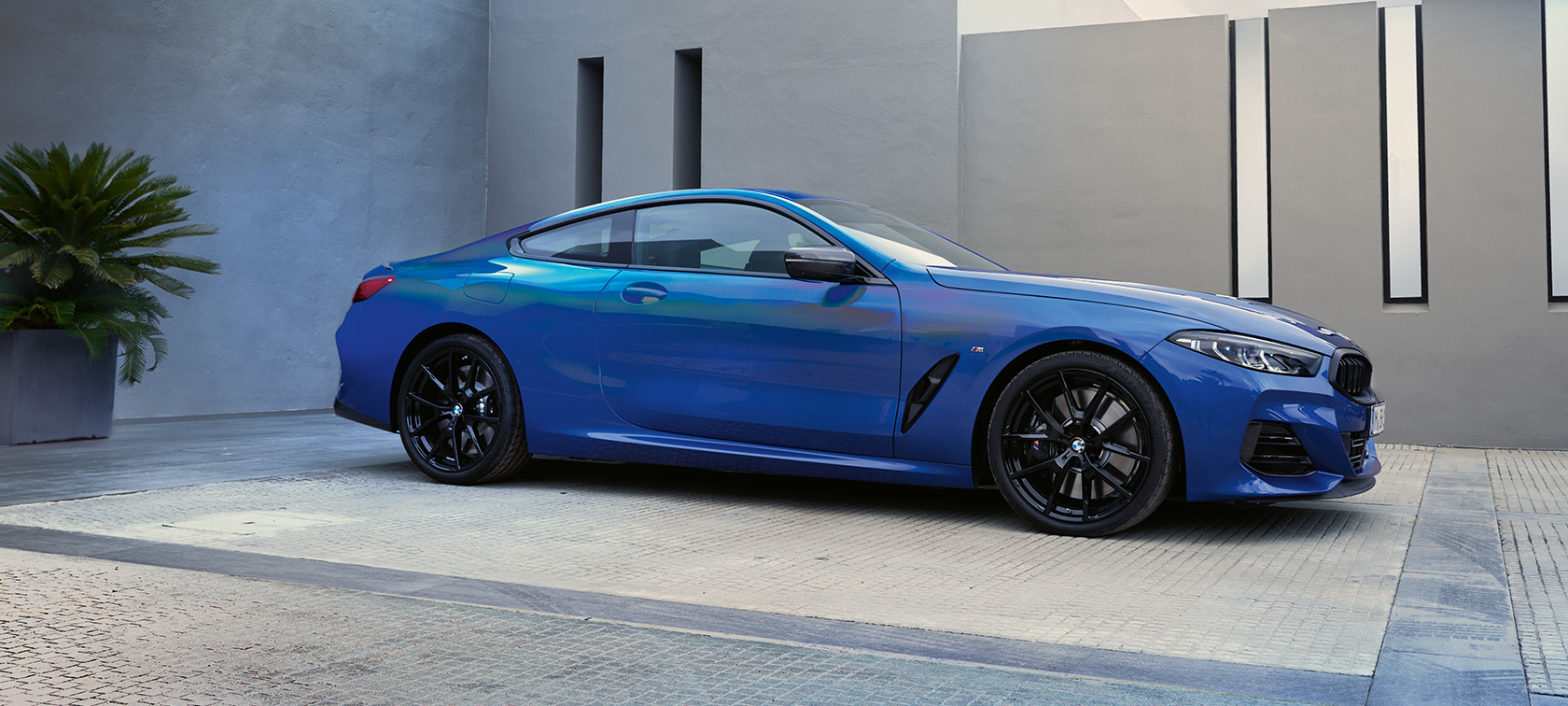 Передня частина BMW M850i xDrive Coupe G15 LCI Facelift 2022 BMW xDrive Coupe G15 LCI Facelift 2022 M у кольорі Portimao Blue metallic, вигляд збоку на фоні стіни будинку