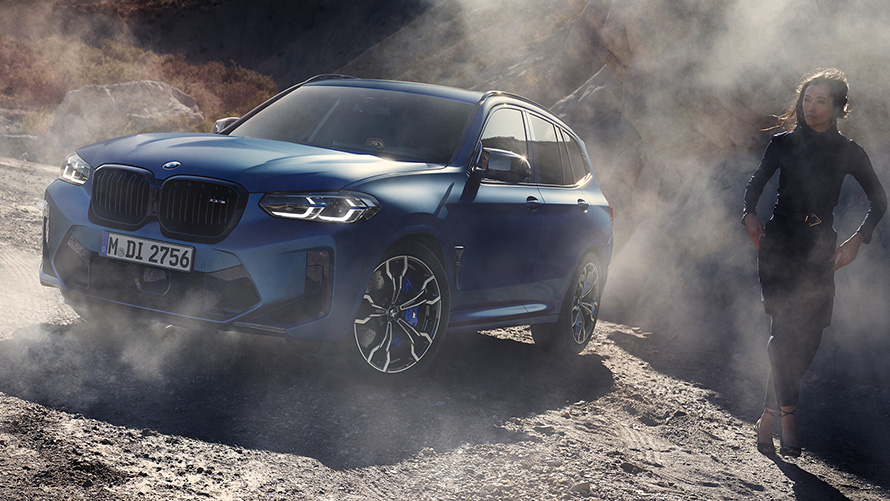 BMW X3 M Competition F97 LCI 2021, вид спереду в три чверті на кам'янистій місцевості з жінкою BMW X3 M Competition F97 LCI Facelift 2021 в кольорі Frozen Marina Bay Blue металік, вид спереду в три чверті на кам'янистій місцевості з жінкою