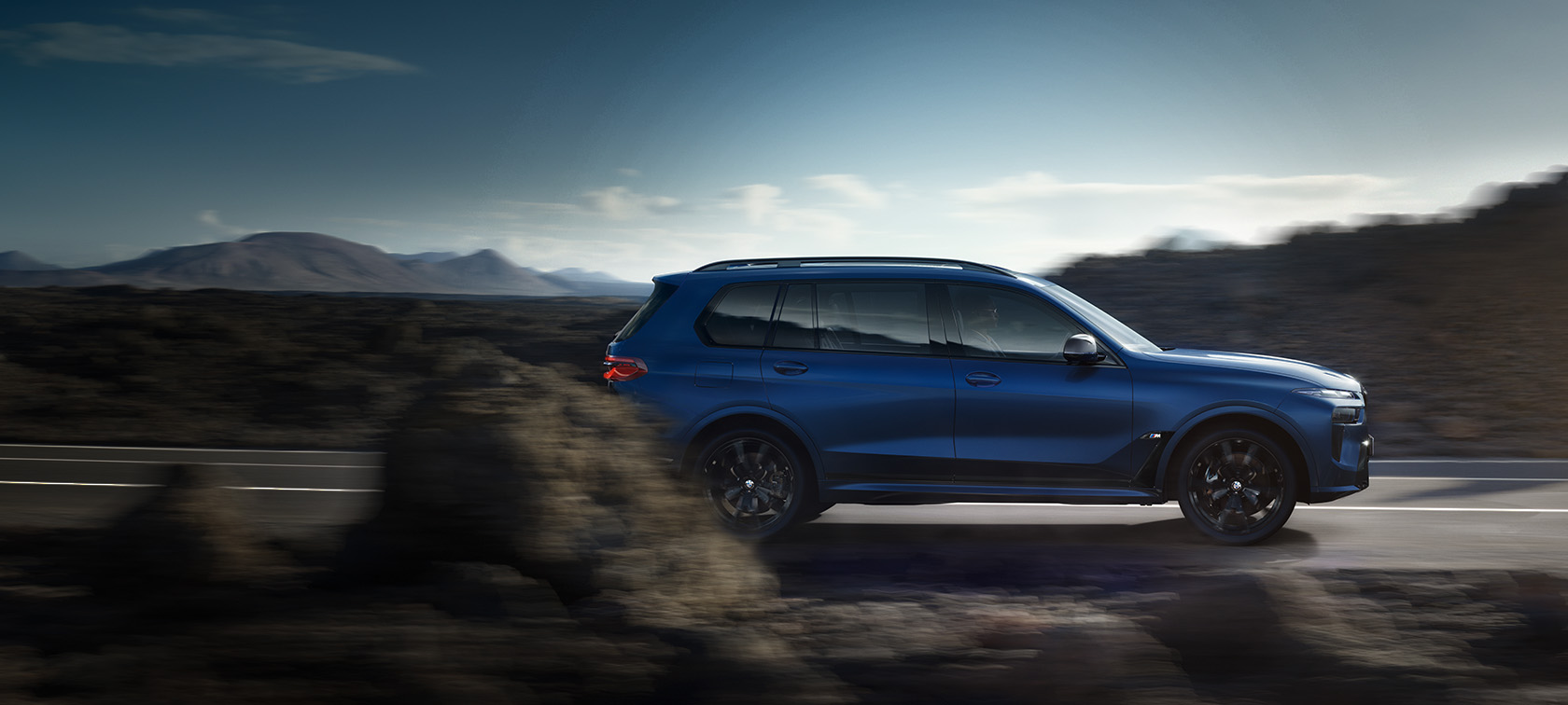 BWM X7 M60i xDrive G07 LCI Frozen Marina Bay Blue вид збоку водіння вулицею BWM X7 M60i xDrive G07 LCI Frozen Marina Bay Blue вид збоку водіння вулицею перед гірським пейзажем